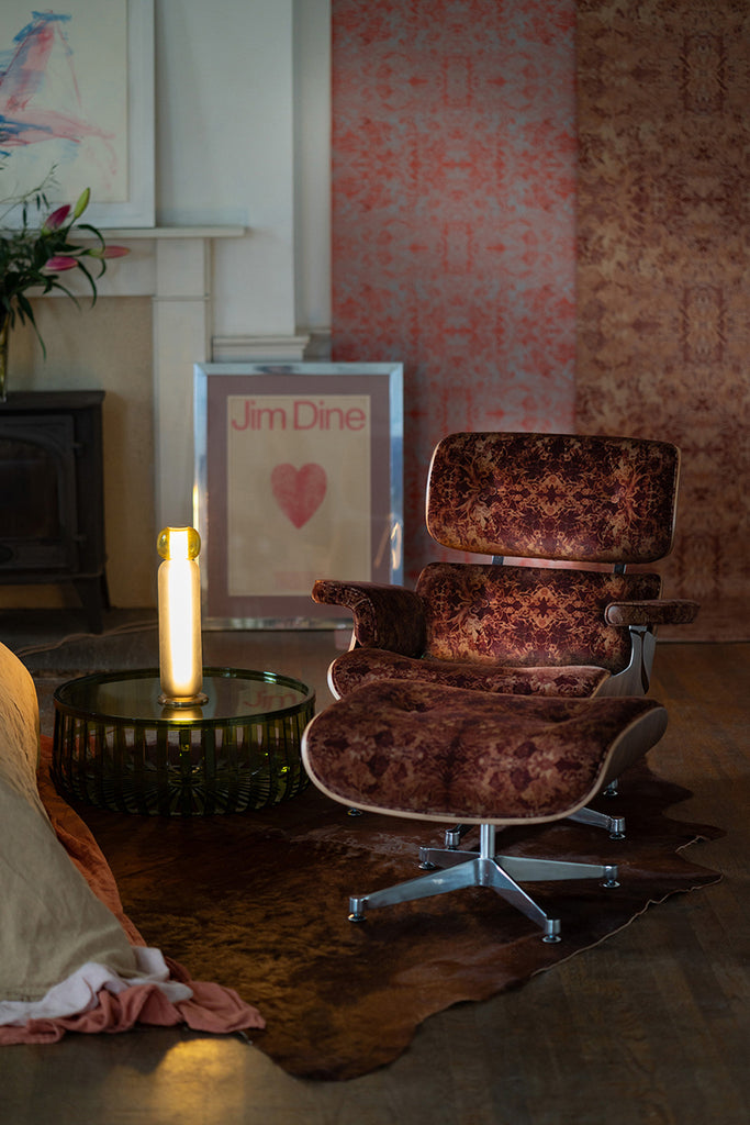 Marlene cotton velvet from Blackpop, this brown patterned velvet covers a mid century armchair and footstool. With a blue pillow in a room with patterned wallpaper and a lit candle.