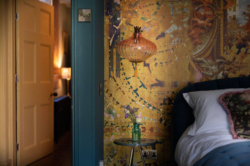 Hampton Gold wallpaper mural styled behind a velvet headboard and white bedding with glass golden pendant light. 