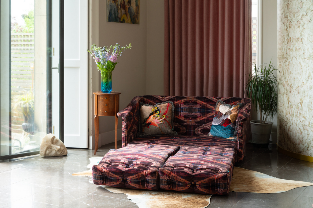 Tartan patterned velvet sofa in a living room with a side table, 70’s house plants, and draped pink curtains.