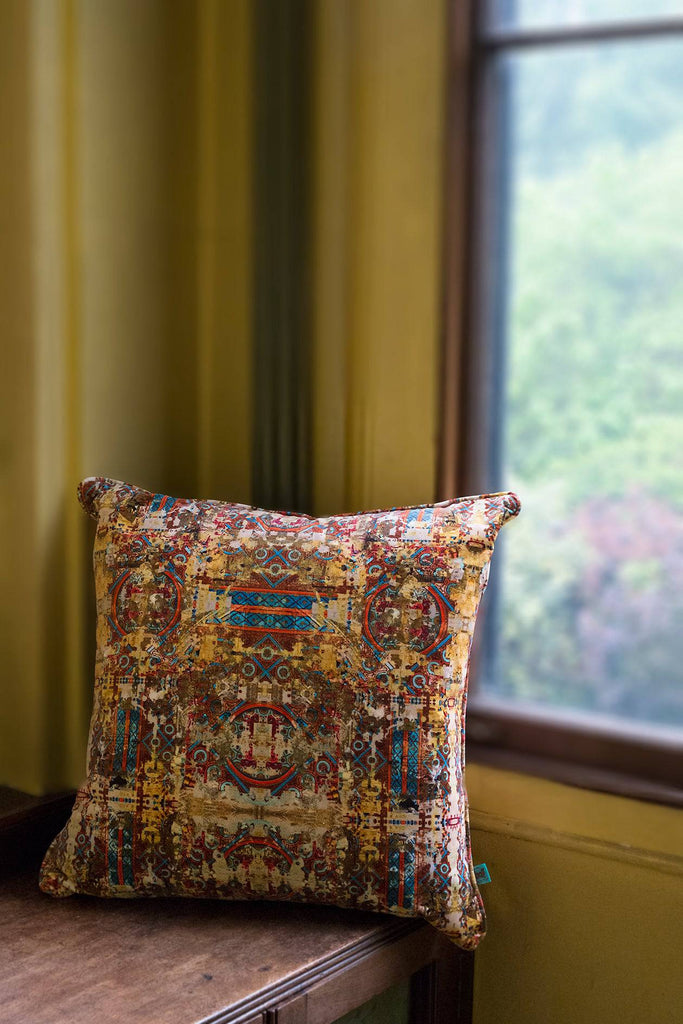 ‘Fresco’ velvet square cushion featuring yellow, red and blue geometric patterns placed on a wooden bench in a yellow interior.